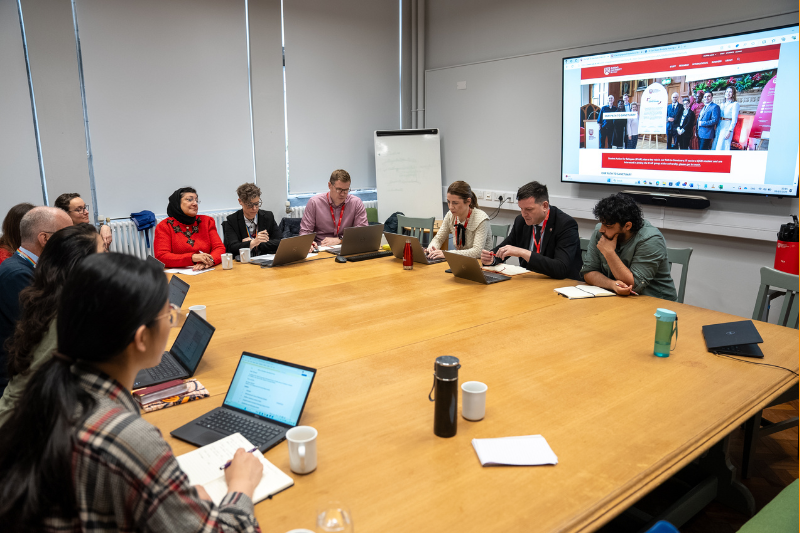 Diverse group of people sitting around a table; behind, screen with Queen's University of Sanctuary webpage